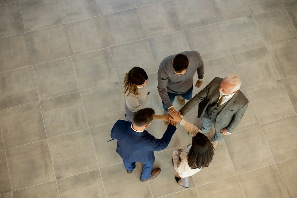 Group of young and senior business people come together in an office hallway, standing in a circle with their hands joined. They are dressed professionally, reflecting their business acumen and status. Their teamwork and collaboration symbolize the unity and strength of the company, as they work towards common goals and successes.